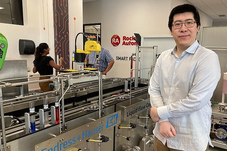 A man wearing a white shirt and standing in a manufacturing environment is smiling as he looks at the camera.