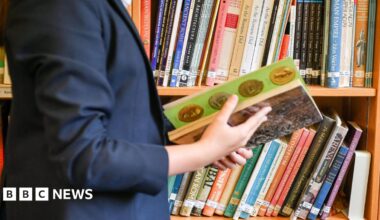 A child wearing a navy blue school blazer looks through a history book in a library. They are stood in front of a bookshelf which has dozens of other history books on it.