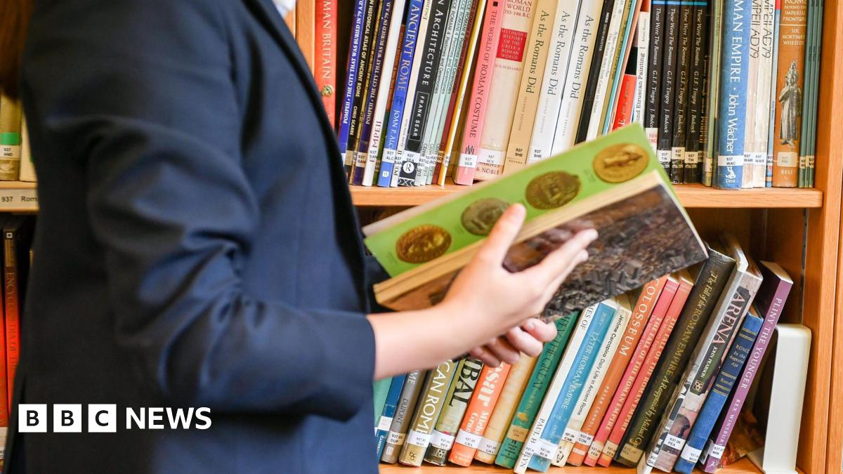 A child wearing a navy blue school blazer looks through a history book in a library. They are stood in front of a bookshelf which has dozens of other history books on it.