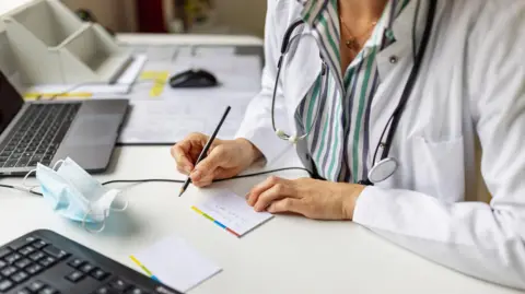 Getty Images A generic image of a caucasian female GP in a white lab coat and striped purple and green shirt writing at a desk. She has a stethescope around her neck. 