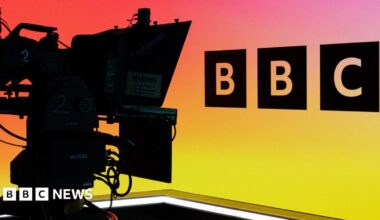 A camera operator in a BBC studio, showing the BBC logo against a multi-coloured backdrop