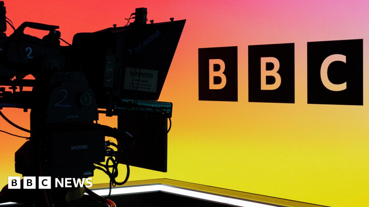 A camera operator in a BBC studio, showing the BBC logo against a multi-coloured backdrop