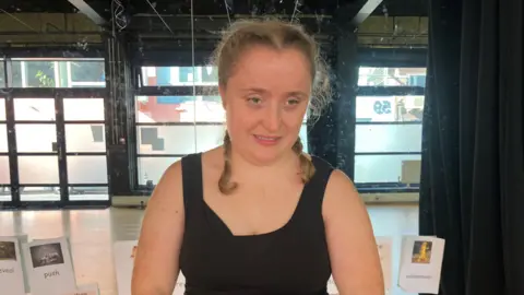 Leanne Rinne / BBC A woman wearing a black sleeveless t-shirt. She is stood in front of a mirrored wall with pictures attached to it.