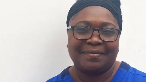 University Hospitals of Northamptonshire A head and shoulders shot of Eunice Vial. She is wearing a blue NHS branded gown and glasses.