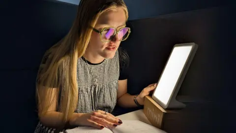 BBC A young woman with long blond hair wearing glasses, a black T-shirt, and a black-and-white checkered dress. She is sitting at a desk or table and reading a book. Next to her is a bright, rectangular light therapy lamp. The light is illuminating her face and the book she is reading. Her nails are painted red with white polka dots.