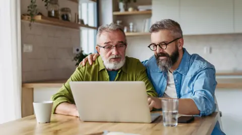 Getty Images An older man with glasses wearing a green shirt next to a younger man with a blue shirt sat in front of a laptop. 