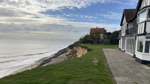 Richard Daniel/BBC A heavily eroded coastline encroaches on a white house. The sea is on the left of the image and another house is in the distance.