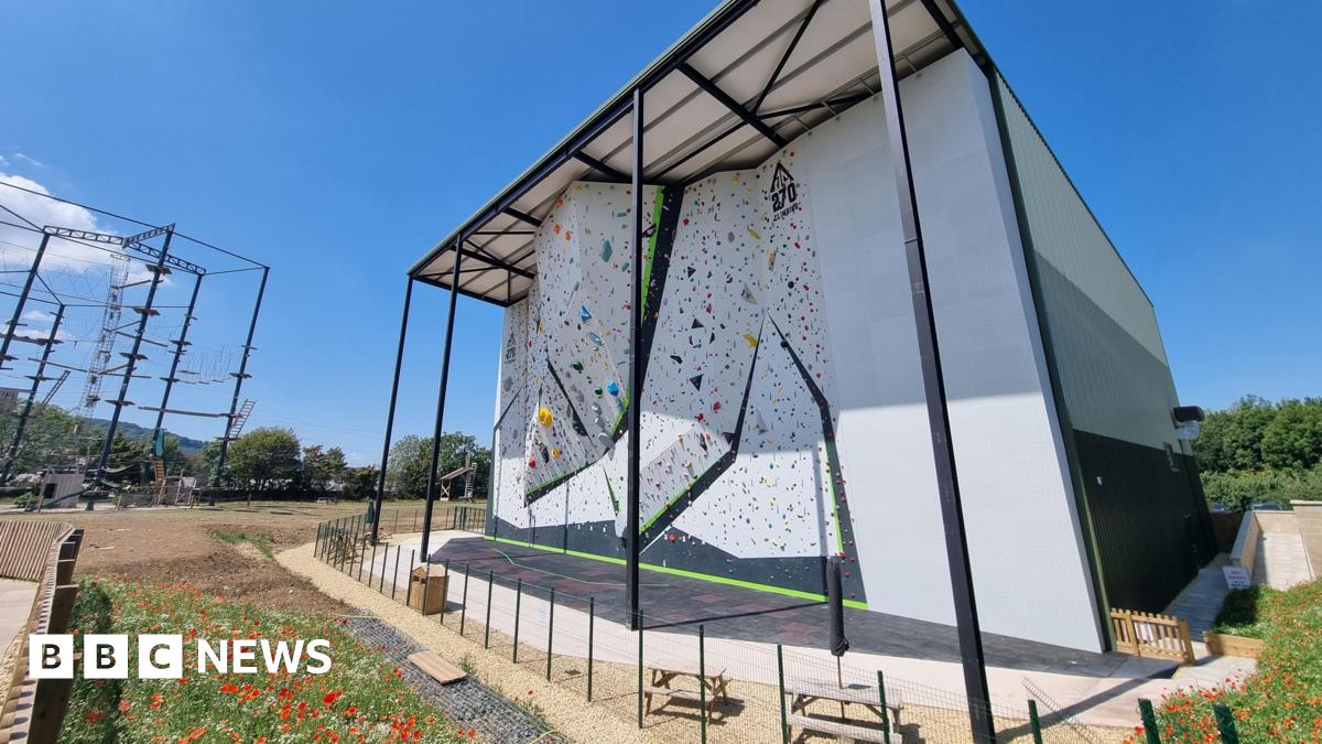 In bright sunshine and near cloudless skies, a giant climbing wall towers over the surrounding area. Several overhangs protrude out from the main structure.