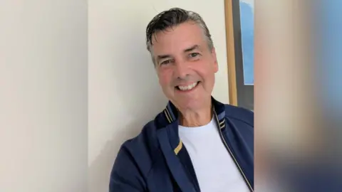 Paddy Lowry has salt and pepper hair parted to the side and is smiling towards the camera. He is wearing a blue jacket and white t shirt