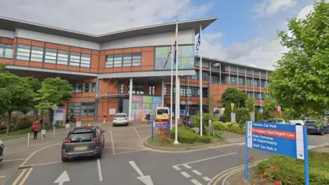 Google Front view of Princess Royal University Hospital showing two cars and an ambulance parked and driving outside it. To the front right of the photo there is a sign showing the way to the car park, emergency department, urgent care and main entrance.