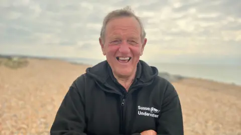 Fiona Irving / BBC A man wearing a black coat stood on a pebble beach with his arms crossed. His coat has white righting on the right breast, reading "Sussex Underwater".