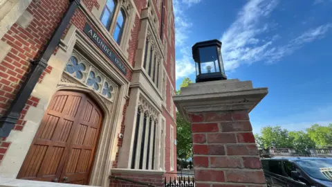 Stuart Woodward/BBC Arundel House is a red brick building with sandstone surrounding the doors and windows. There is a pair of closed wooden double doors at the front of the building with the words Arundel House on a black sign above the doorframe. There is a brick pillar with a black lantern on top at the front of the building, and there is a car parked next to the pillar. There are green trees in the distance and blue sky above.