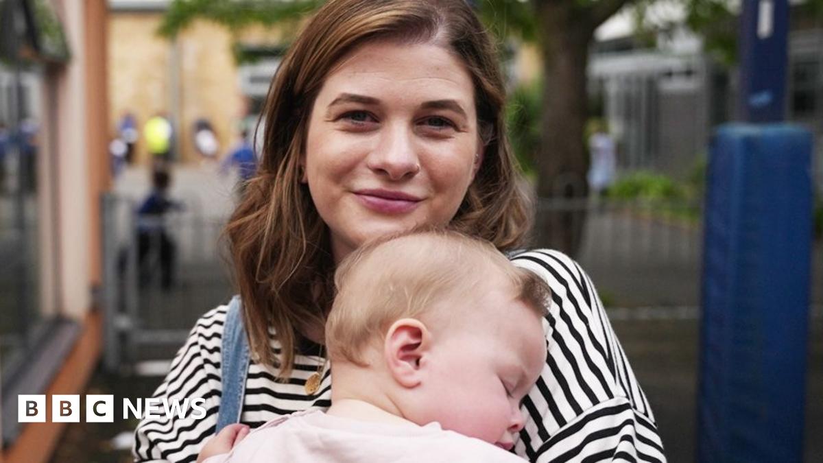 A portrait of Holly and her young daughter Ettie. Holly is a young woman aged 30 with long brown hair and  wearing a striped blue top. She is carrying her baby Elly who is asleep in her arms and wearing a very light pink top with long sleeves.