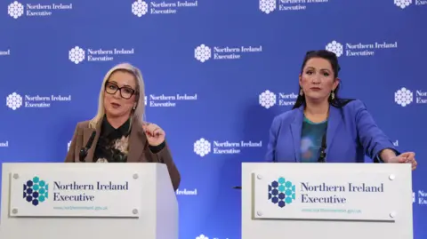 PA Media Two women stand at lecterns in front of a blue background. The blue background has the words 'Northern Ireland Executive' written on it multiple times. The same words are written on both lecterns. The woman on the left has blonde hair and is wearing glasses. She is wearing a dark shirt and a brown blazer. The woman on the right is wearing a green top and a lilac blazer. She has dark hair. 