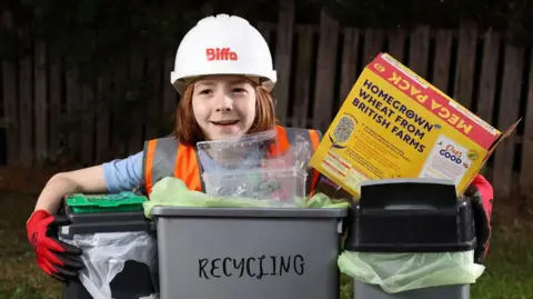 Glen Minikin Tommy crouched behind three recycling bins holding a cardboard cereal box. He is wearing a high visibility orange bib, red gloves and a white hard hat with Biffa written on it in red writing