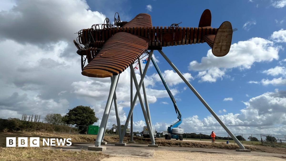 A life-size steel brown sculpture of a Lancaster bomber in a flying motion and tilted to the side. It is held up high by several posts and construction vehicles and a person in orange hi-vis can be seen next to it.