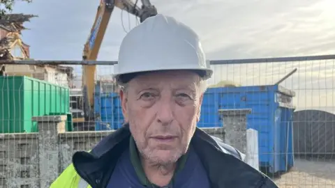 Richard Daniel/BBC A head and shoulders image of Bill Seal. He is wearing a hard hat and standing in front of a metal fence, behind which is a large blue tip and a yellow crane. He is looking directly into the camera.