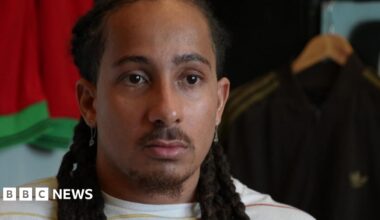 A head-and-shoulders image of Tyler Attwood who is looking serious. He has long dark brown hair styled in dreadlocks. There are sports tops on hangers behind him.