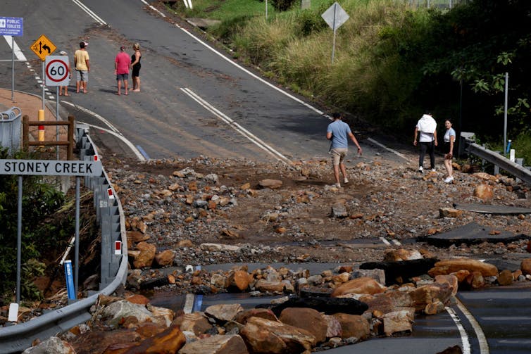 people walk and ride over a debris-covered road in aftermath of flooding.