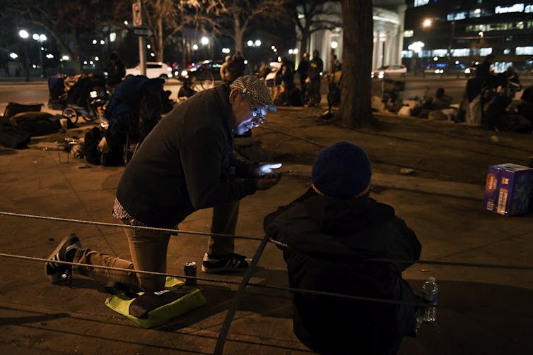 A man kneels with a cell phone in his hand next to a person who is sitting and holding a cane.