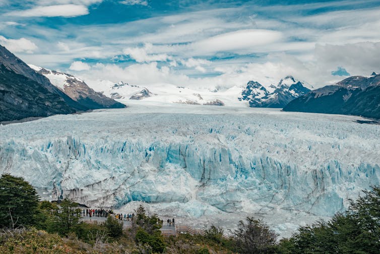 People walk along a path near the face of a large swath of white and blue ice.
