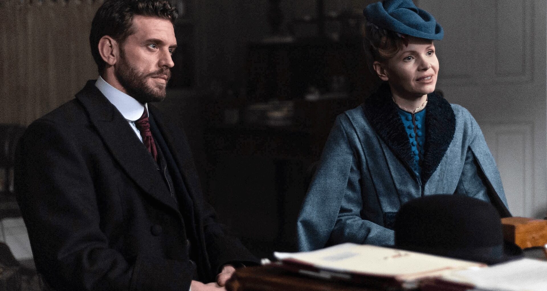 Color image of a scene from Miss Scarlet Season 6 on MASTERPIECE on PBS. Actor Tom Durant-Pritchard as DI Alexander Black is seated next to Kate Phillips as Eliza Scarlet behind a desk.