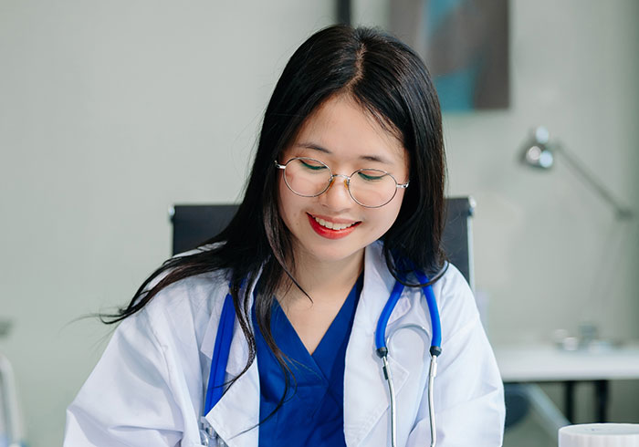 Female doctor wearing a white coat and stethoscope, smiling while reviewing medical information in a clinical setting.