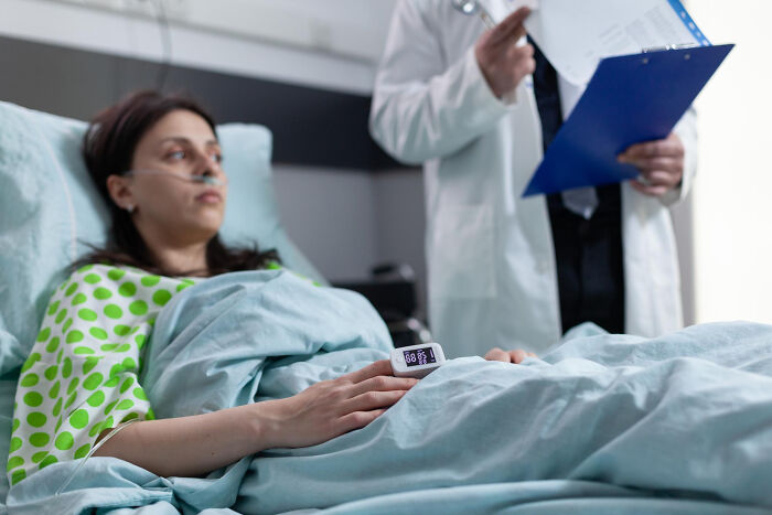 Patient lying in hospital bed with oxygen tube and pulse monitor while doctor reviews medical reports nearby.