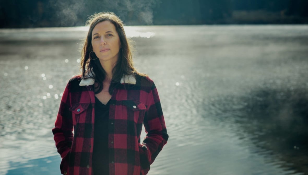 A woman stands in front of a lake, dressed in a red and black plaid jacket. 