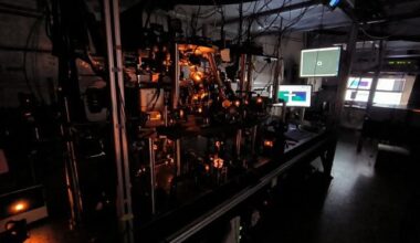 Photo of a darkened optics laboratory with screens and a vacuum system. The scene is bathed in orange light from the lasers used