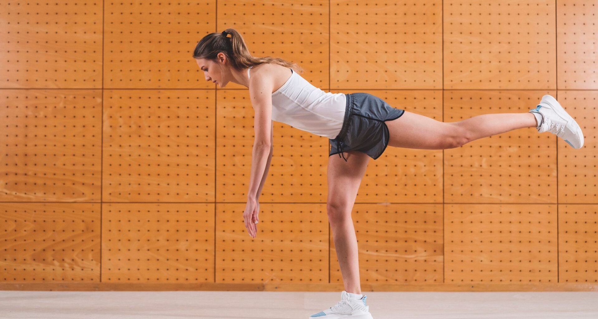 woman in a white vest and grey shorts against an orange background performing a single leg balance side on to the camera.