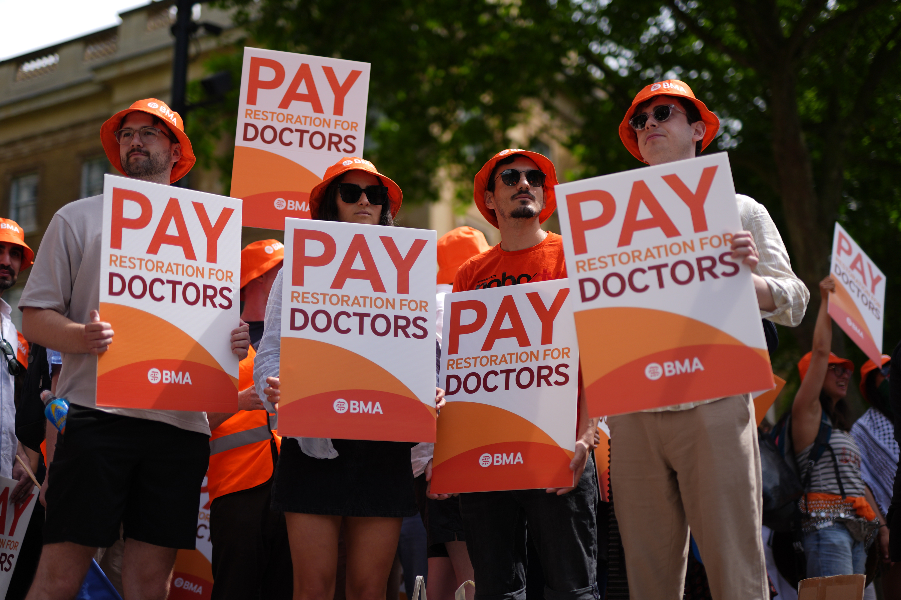 Protesting junior doctors hold signs that read, "Pay Restoration for Doctors" and wear orange bucket hats.