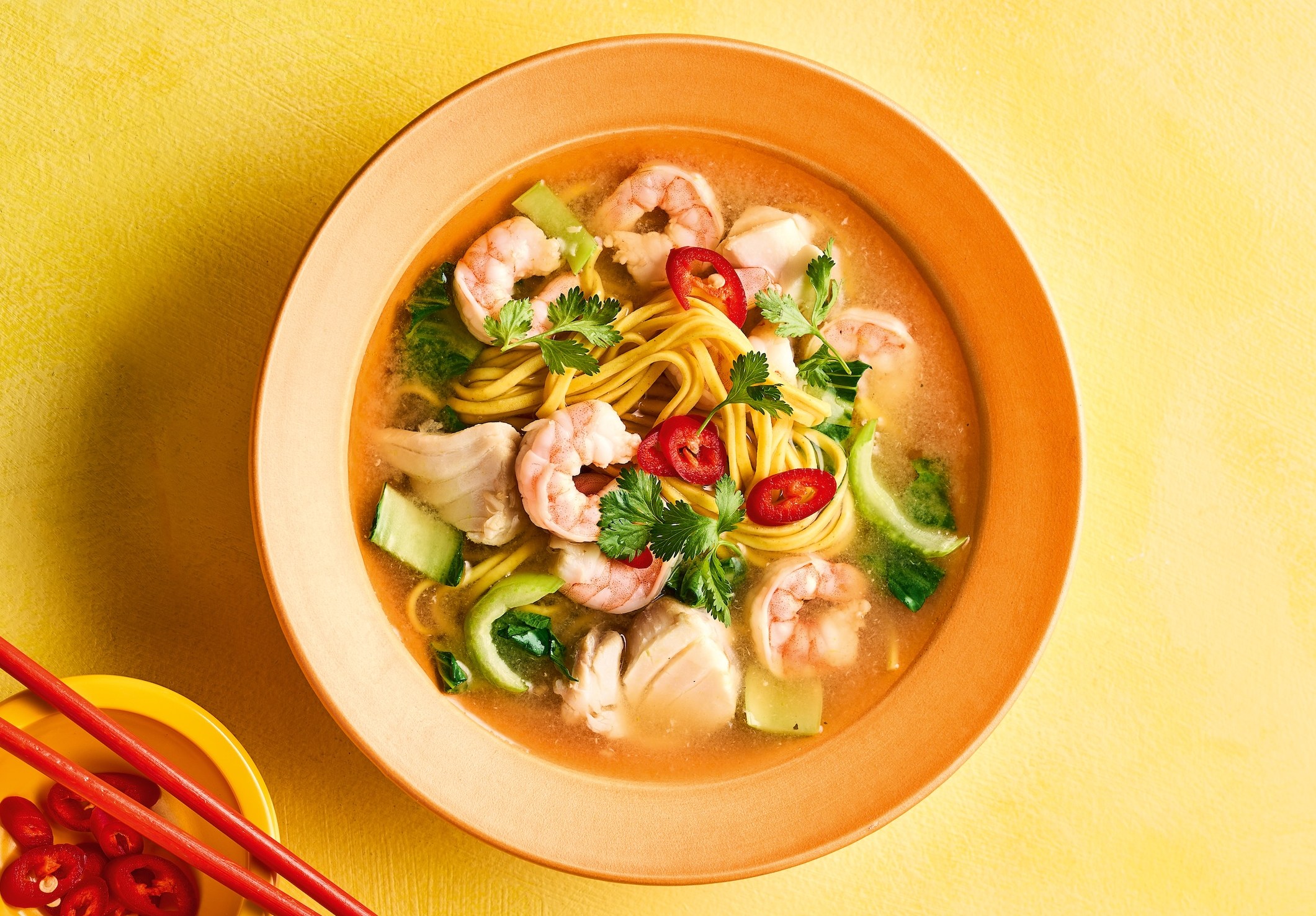 A bowl of seafood broth with noodles, shrimp, and vegetables, with red chopsticks and sliced chilies in a small bowl next to it.
