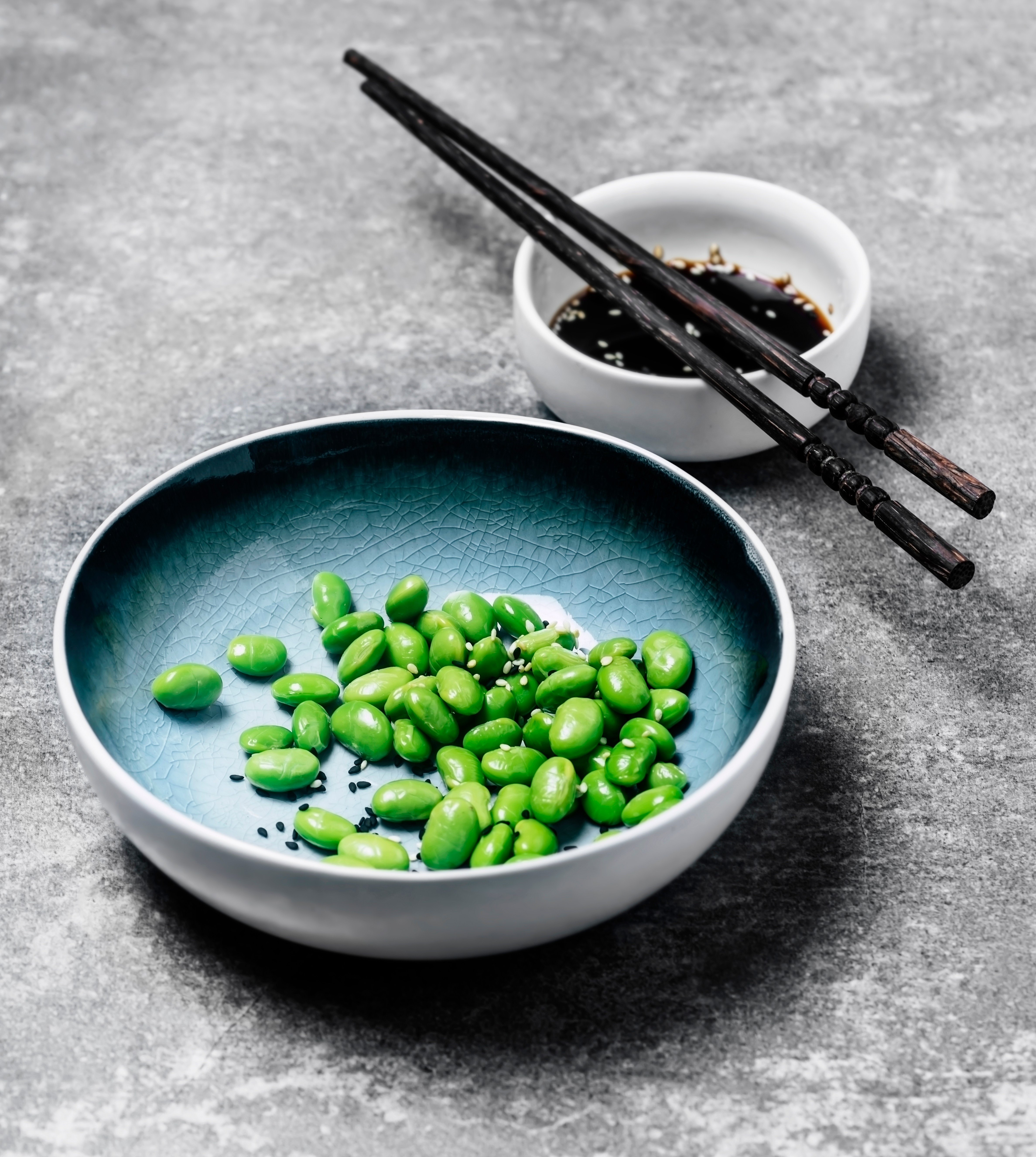 A bowl of edamame with sesame seeds and soy sauce on a gray background.
