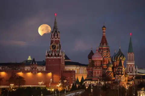 Marina Lystseva / Reuters The Moon rises behind pointy Cathedral buildings with architectural detail and streetlights in the foreground in Moscow on Wednesday. The domes and turrets of the cathedral are richly decorated with stripes and zigzags.