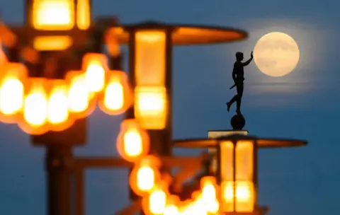 Julian Stratenschulte / DPA via AP Bright orange lights are seen in the foreground next to a statue of someone with their hand out, which the photographer has lined up to make it look like the statue is touching the Moon, at dusk in Hannover on Wednesday.