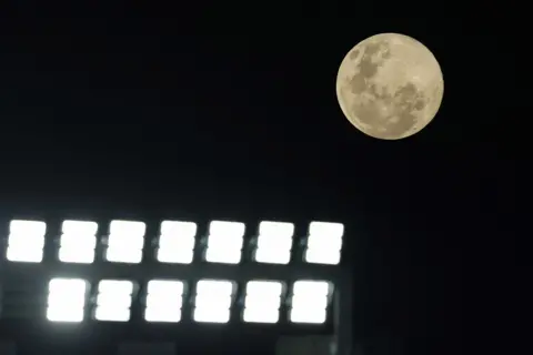 Michael Bradley / AFP via Getty Images The Moon is seen clearly on the right of frame against a dark starless sky, while floodlights glare in the foreground, in Auckland on Wednesday.