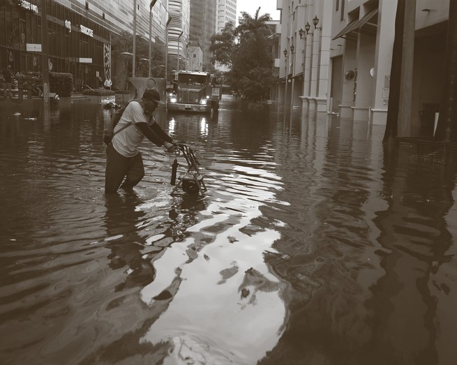 black-and-white photo of man pushing a walker through knee-deep standing water on a downtown street with tall buildings and a tow truck