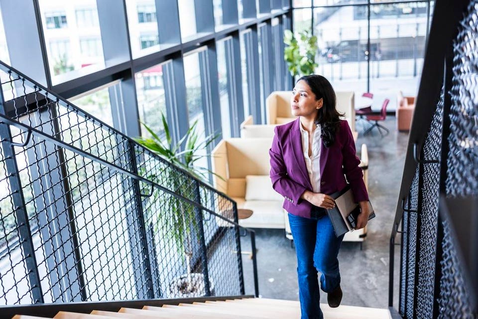 Confident businesswoman walking up modern office staircase and looking out window showing performance under pressure