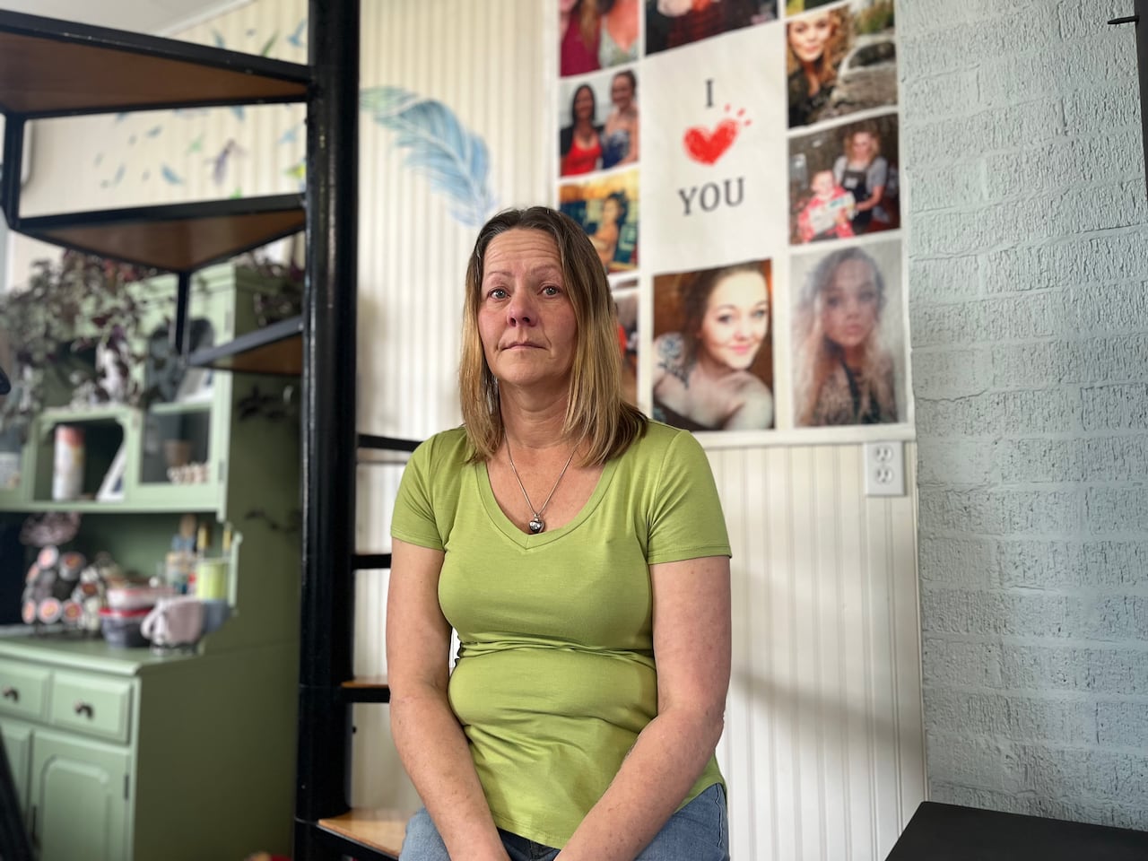 Woman in green T-shirt stands before a wall of photos of a much younger woman.