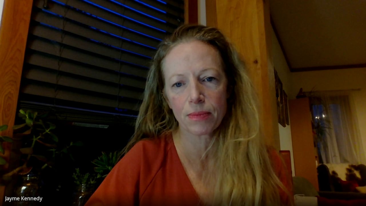 A blond woman speaks in a wood-panelled room.