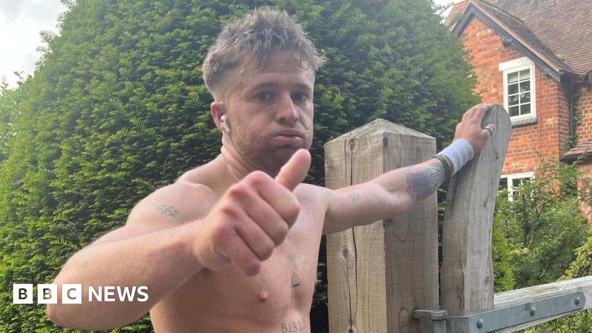 Tommy, a young man, has his top off and is doing a thumbs up at the camera with a tired look on his face. He is resting his other hand on a wooden gate by a house. He has several tattoos, including a cross on his sternum.