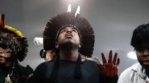 EPA Indigenous people at a protest. Man in centre wears a headpiece made of features and red dye or ink on fingers