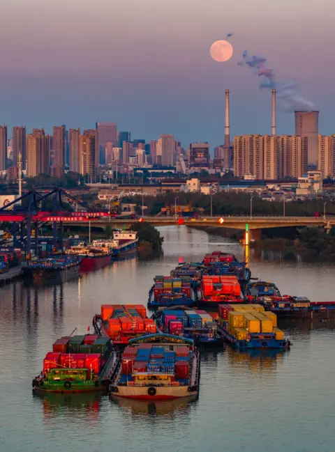 Costfoto/NurPhoto via Getty Images Freight ships are seen moving down the canal in front of a bridge and the city skiyline, with power plant chimneys smoking next to the Moon rising over the city, in Huai'an city on Wednesday.
