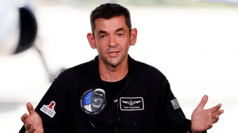 Reuters Jared Isaacman is wearing a black jumpsuit with Polaris patches on the chest. He has short brown hair and is gesturing with both hands as he speaks.
