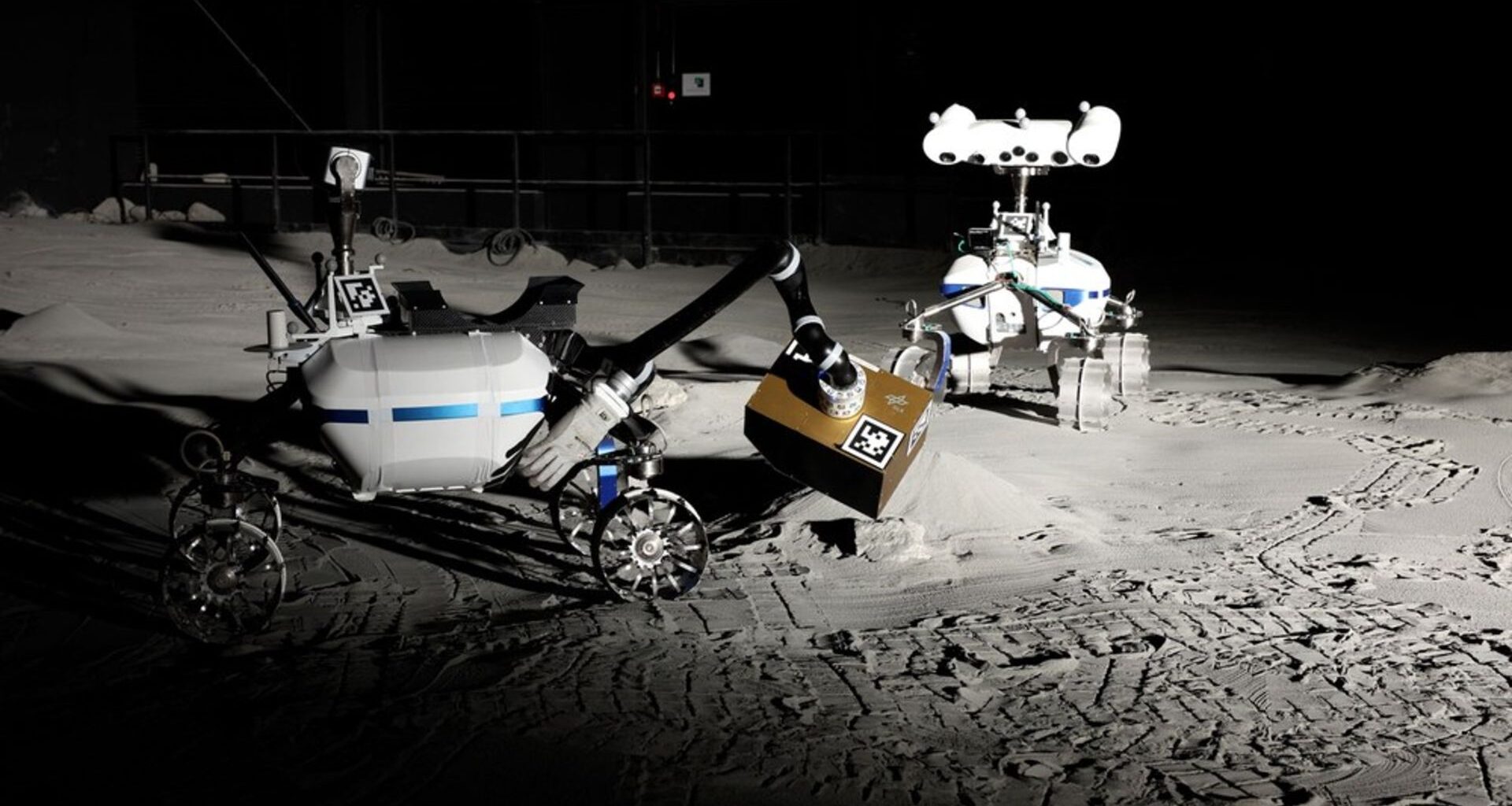 Two white rovers lean over simulated gray lunar dust.