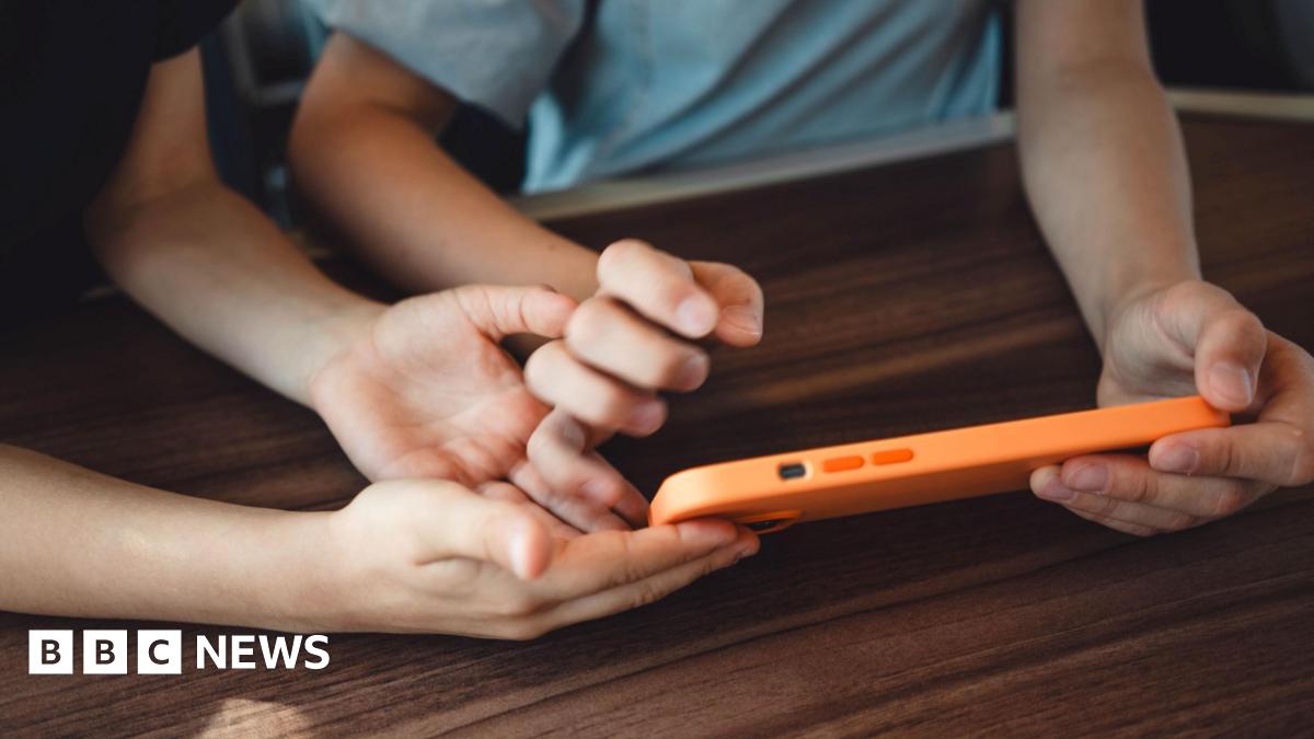 Two children, their faces not visible, hold a smart phone in an orange case on a brown table top.