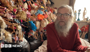 David is sitting in front of a huge pile of Barbie dolls sticking out of several bin bags. He has long grey hair and a beard and glasses. He is wearing a red jumper.