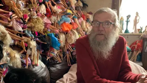BBC David is sitting in front of a huge pile of Barbie dolls sticking out of several bin bags. He has long grey hair and a beard and glasses. He is wearing a red jumper.