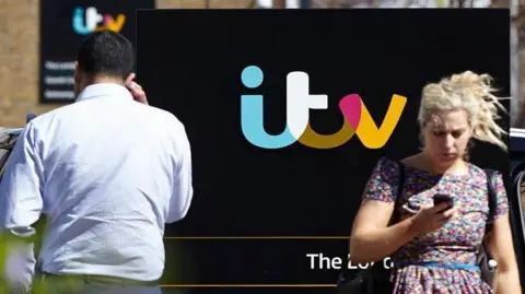Alamy A blonde woman in a floral dress walks along a street while looking at her phone. A man in a white shirt, with his back to the camera, walks in the opposite direction with his back to the camera. A black sign with the multi-coloured ITV logo can be seen in the background. 